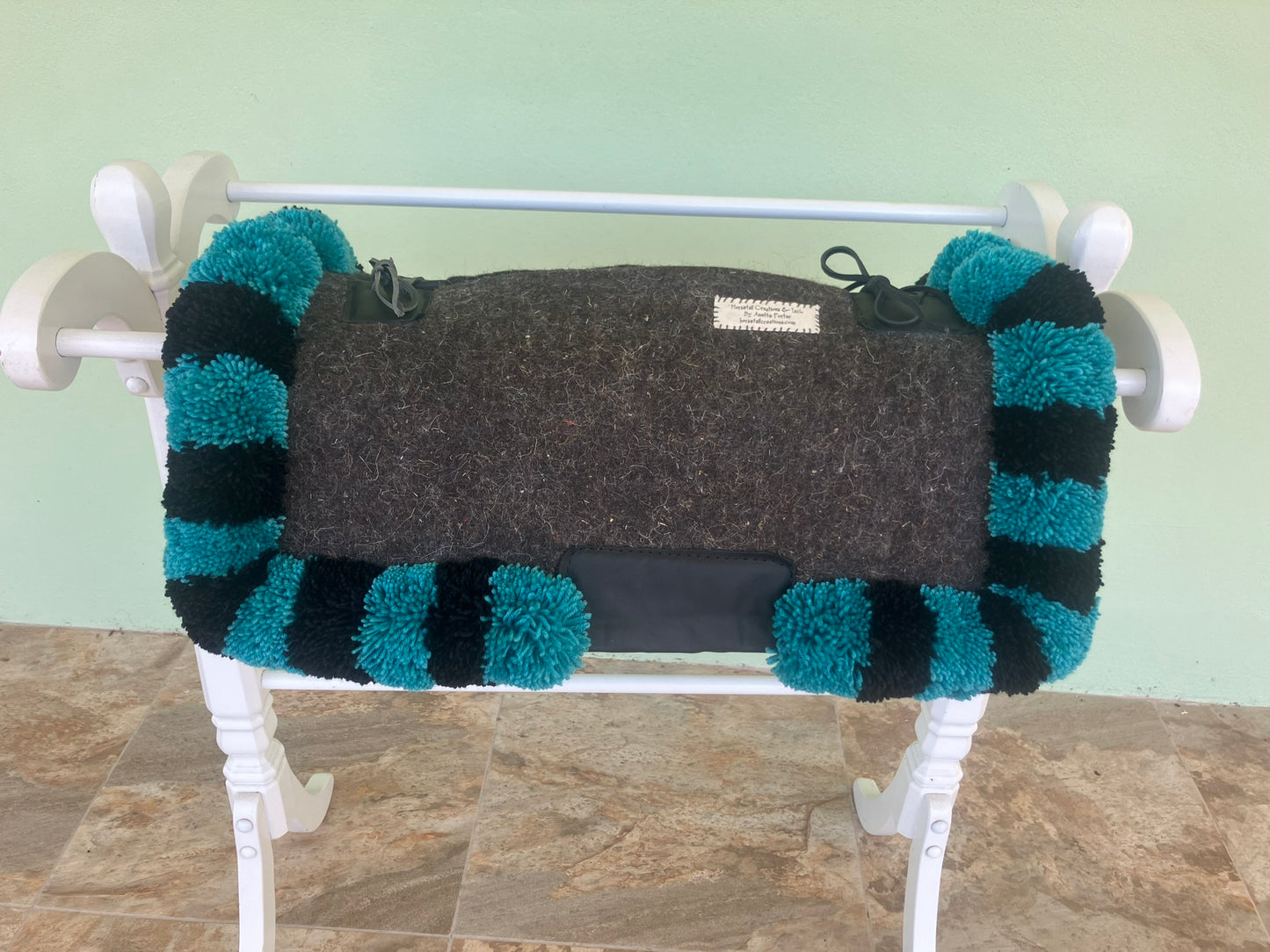 Gray saddle pad with teal and black striped edges draped over a white rack against a light green wall.